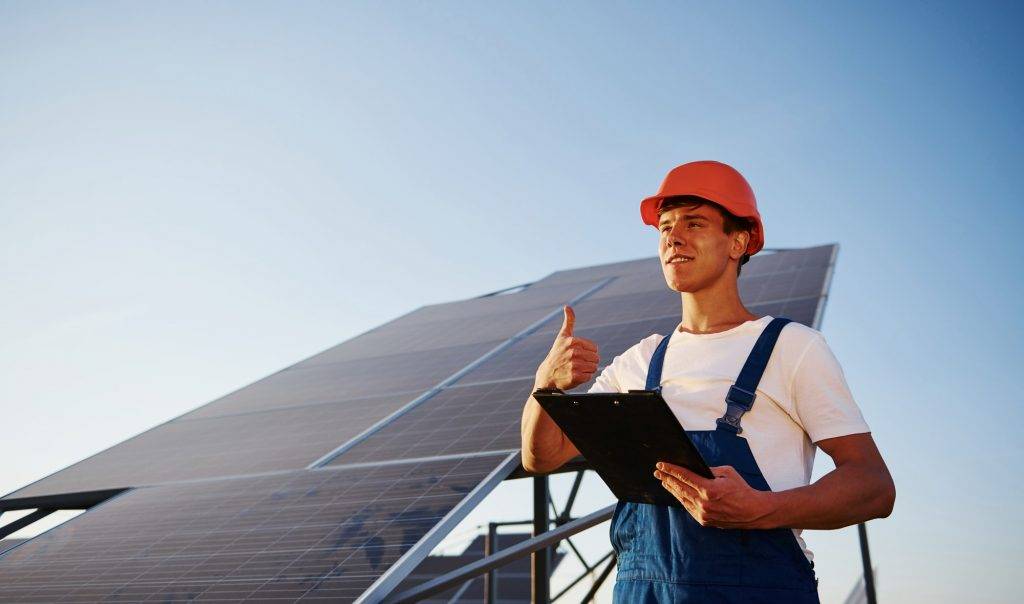 shows thumb up male worker in blue uniform outdoors with solar batteries at sunny day